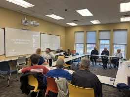 photo of about 8 adults sitting at tables arranged in a square and looking a screen with the phrase "I've got so much honey the bees envy me" projected on it.